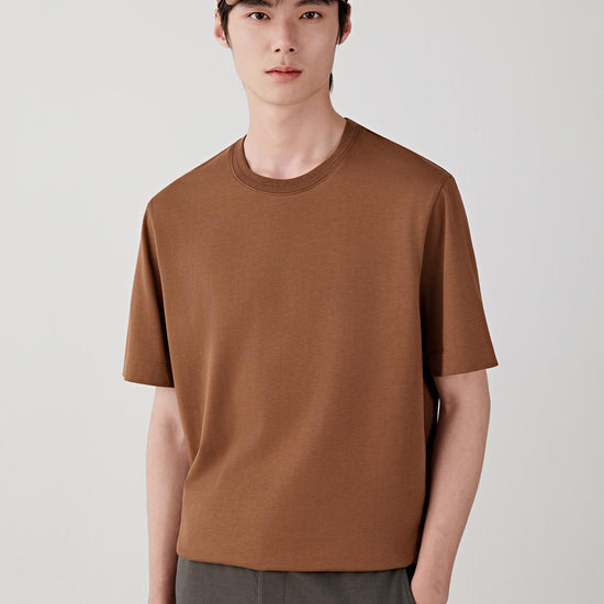 Man wearing a chestnut brown cotton jersey T-shirt with a relaxed straight cut and round neckline, styled with dark trousers and a cap. He moves through front, side and back views, showing the lightweight, breathable cotton jersey and its smooth, even drape. The clean neckline, short sleeves and minimal construction remain consistent, while the fabric follows the body naturally, emphasizing comfort, ease of movement and a simple, balanced everyday fit.
