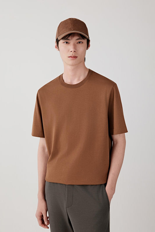 Man wearing a chestnut brown cotton jersey T-shirt with a relaxed straight cut and round neckline, styled with dark trousers and a cap. He moves through front, side and back views, showing the lightweight, breathable cotton jersey and its smooth, even drape. The clean neckline, short sleeves and minimal construction remain consistent, while the fabric follows the body naturally, emphasizing comfort, ease of movement and a simple, balanced everyday fit.