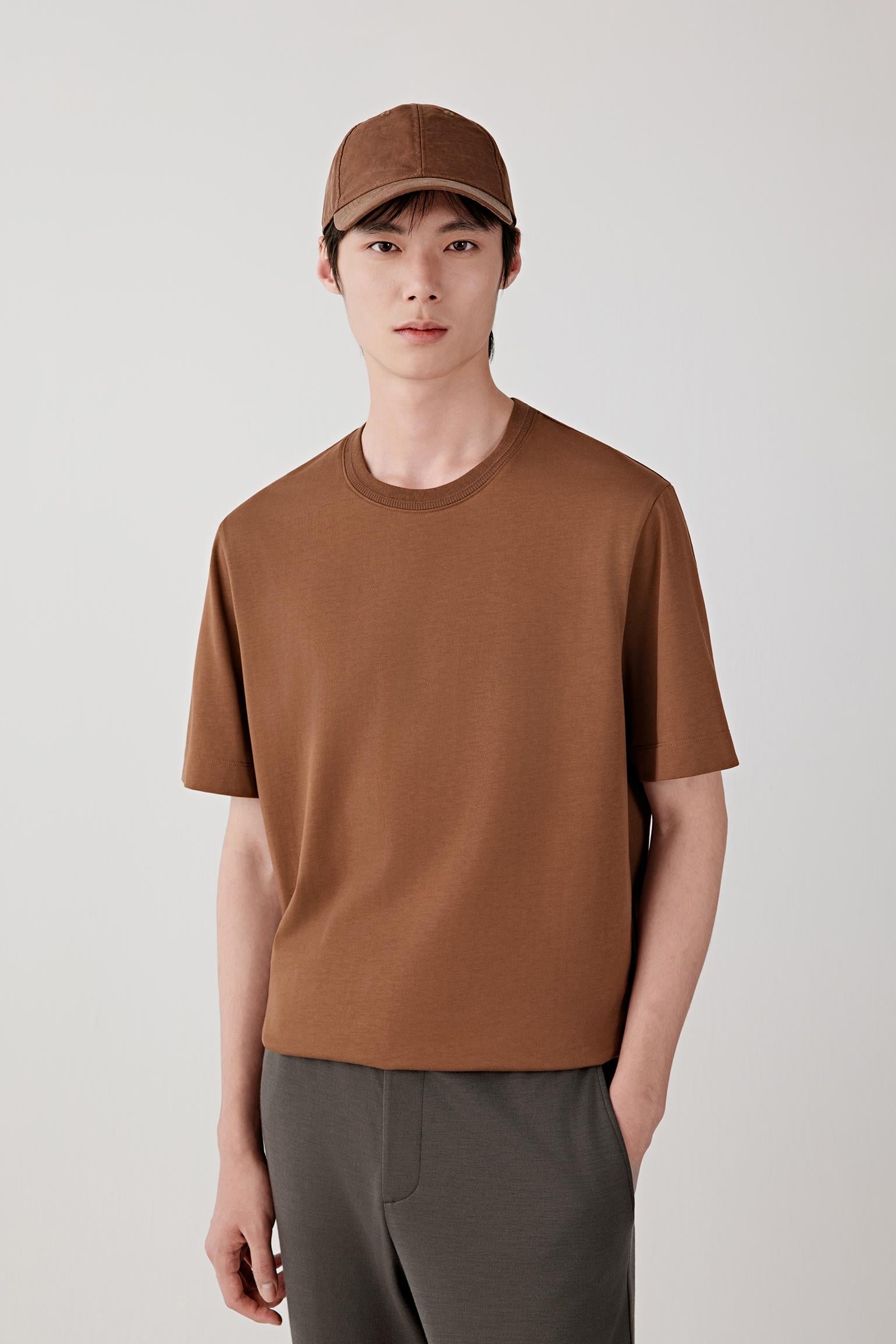 Man wearing a chestnut brown cotton jersey T-shirt with a relaxed straight cut and round neckline, styled with dark trousers and a cap. He moves through front, side and back views, showing the lightweight, breathable cotton jersey and its smooth, even drape. The clean neckline, short sleeves and minimal construction remain consistent, while the fabric follows the body naturally, emphasizing comfort, ease of movement and a simple, balanced everyday fit.