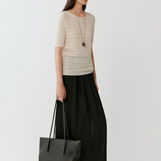 Woman wearing a soft cream silk and linen blend short-sleeved knit top with a relaxed, straight fit, styled with a long black skirt. She moves through front and side views, showing the lightweight open-knit texture, breathable structure and gentle drape. The round neckline and short sleeves remain visible, while the semi-sheer knit allows light through, emphasizing airflow, comfort and a fluid silhouette.