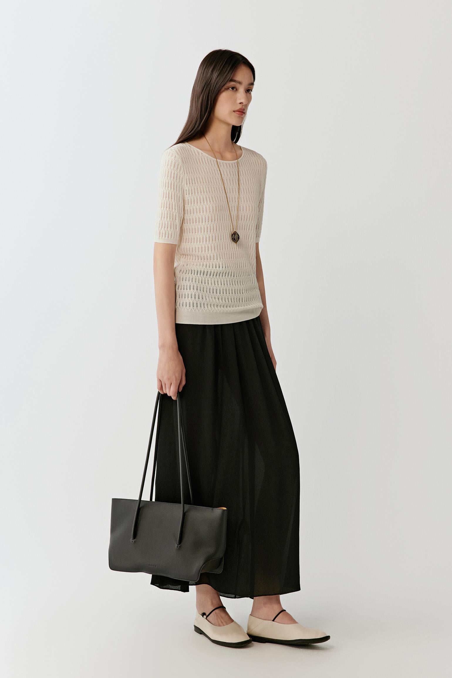 Woman wearing a soft cream silk and linen blend short-sleeved knit top with a relaxed, straight fit, styled with a long black skirt. She moves through front and side views, showing the lightweight open-knit texture, breathable structure and gentle drape. The round neckline and short sleeves remain visible, while the semi-sheer knit allows light through, emphasizing airflow, comfort and a fluid silhouette.