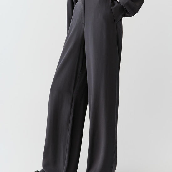 Woman wearing black silk crepe de chine trousers with a fluid, wide-leg cut, styled with a matching shirt. She moves through front and side views, showing the lightweight fabric, smooth drape and slightly matte finish. The high waist and clean lines remain visible, while the trousers fall softly along the leg, emphasizing fluid movement and a relaxed silhouette.