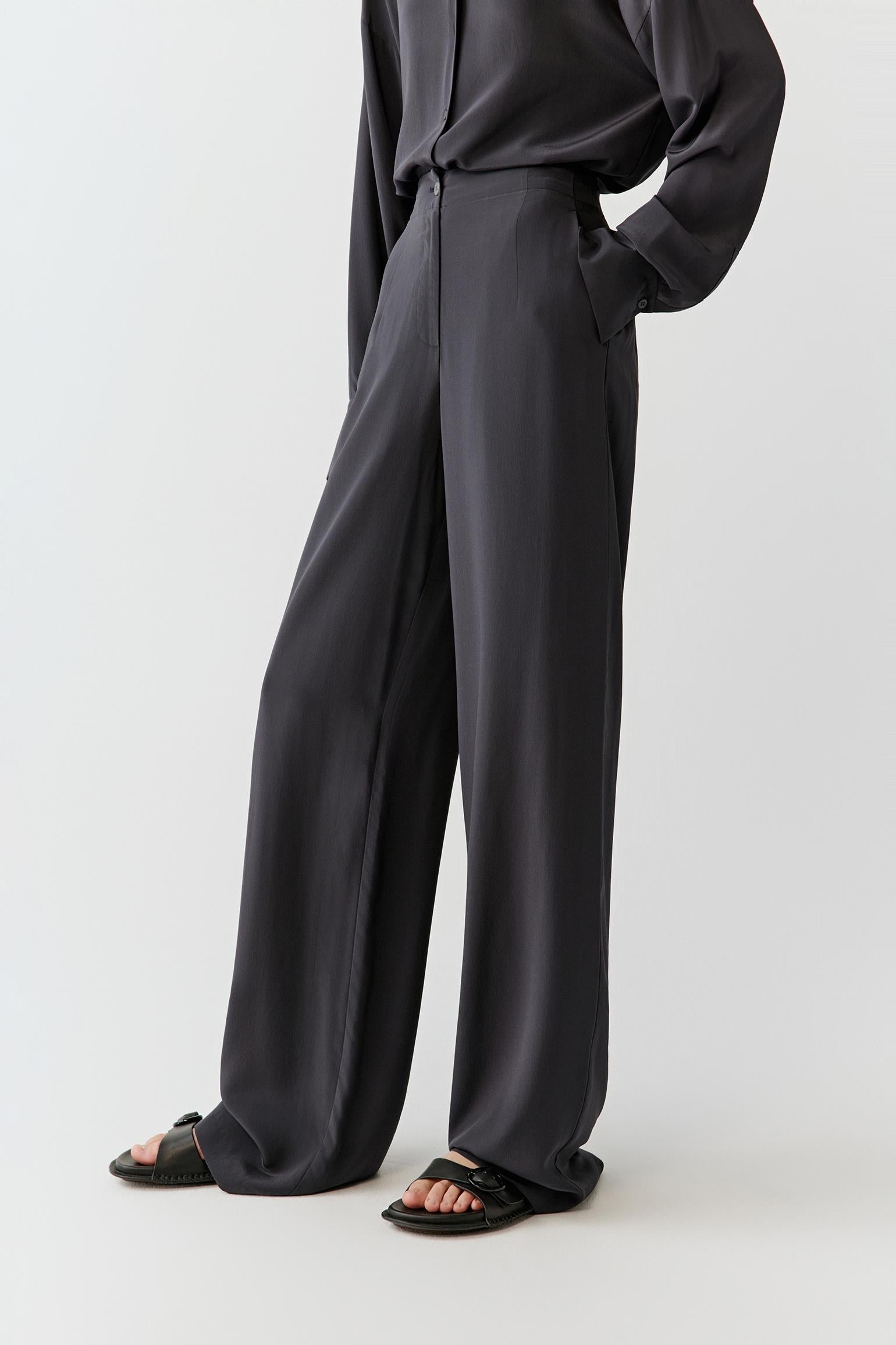Woman wearing black silk crepe de chine trousers with a fluid, wide-leg cut, styled with a matching shirt. She moves through front and side views, showing the lightweight fabric, smooth drape and slightly matte finish. The high waist and clean lines remain visible, while the trousers fall softly along the leg, emphasizing fluid movement and a relaxed silhouette.