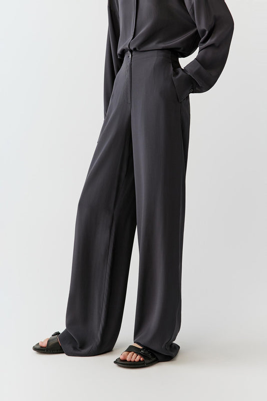 Woman wearing black silk crepe de chine trousers with a fluid, wide-leg cut, styled with a matching shirt. She moves through front and side views, showing the lightweight fabric, smooth drape and slightly matte finish. The high waist and clean lines remain visible, while the trousers fall softly along the leg, emphasizing fluid movement and a relaxed silhouette.