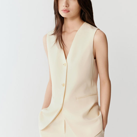 Woman wearing an ivory silk stretch heavy crepe vest jacket with a fluid, tailored fit, styled with matching trousers. She moves through front and side views, showing the smooth fabric, soft structure and clean lines. The V-neckline, tonal button fastening and sleeveless cut remain visible, while the vest falls straight along the torso, emphasizing lightness, ease of movement and a refined silhouette.