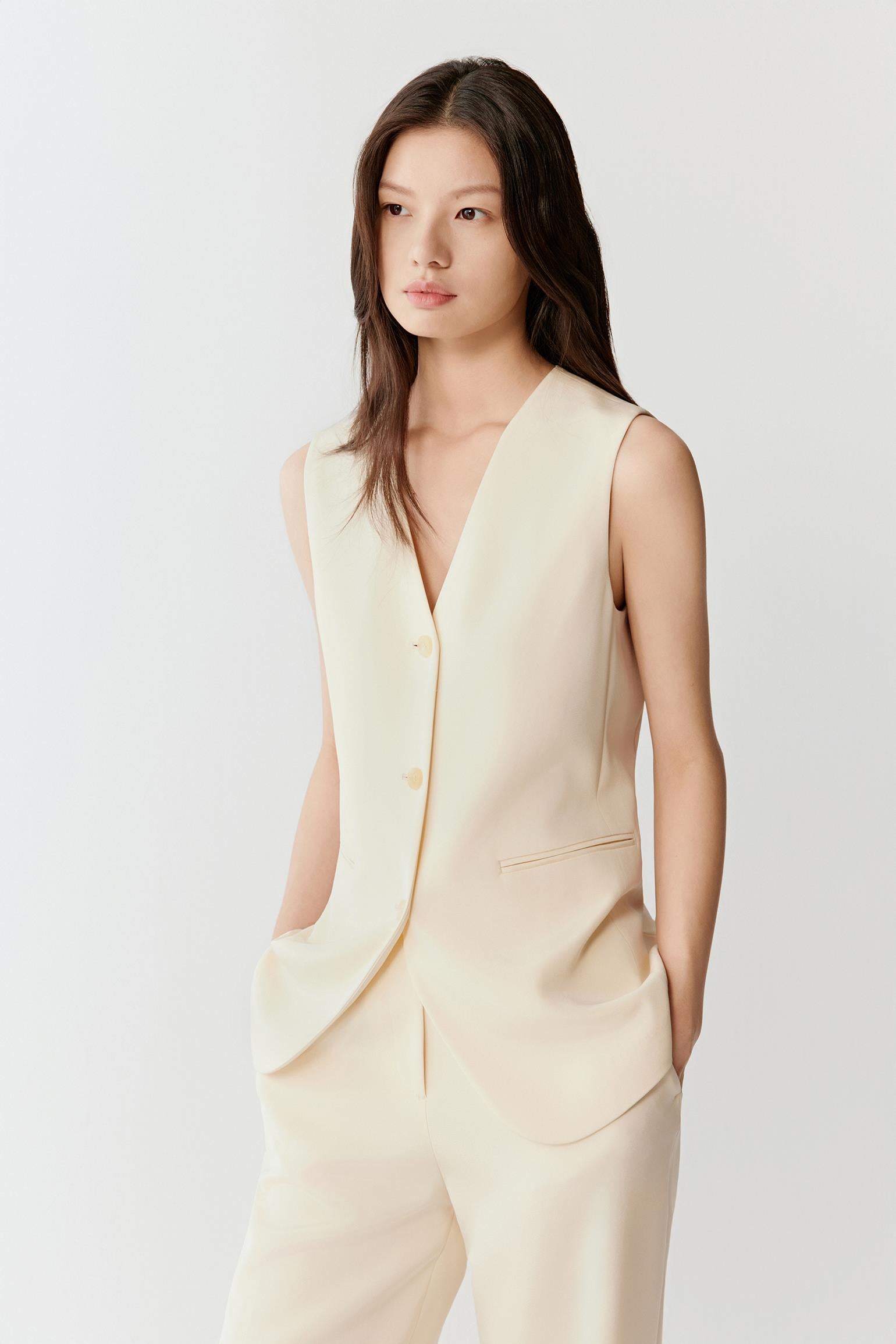 Woman wearing an ivory silk stretch heavy crepe vest jacket with a fluid, tailored fit, styled with matching trousers. She moves through front and side views, showing the smooth fabric, soft structure and clean lines. The V-neckline, tonal button fastening and sleeveless cut remain visible, while the vest falls straight along the torso, emphasizing lightness, ease of movement and a refined silhouette.