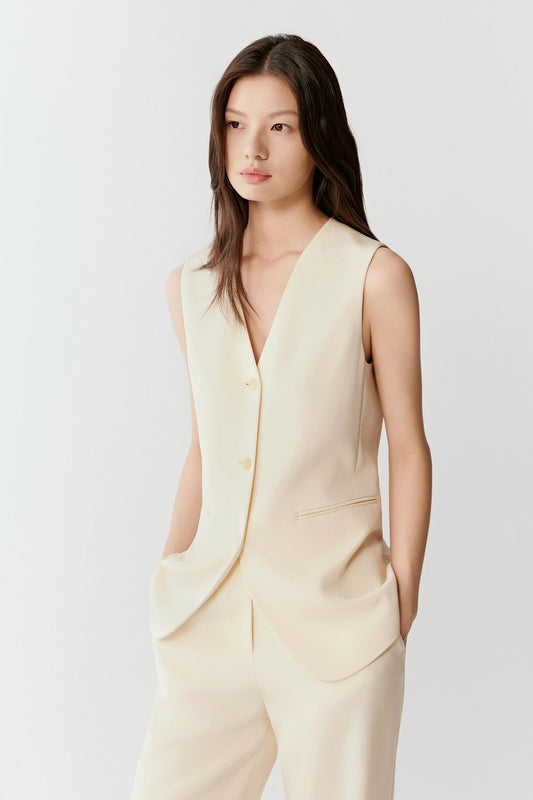 Woman wearing an ivory silk stretch heavy crepe vest jacket with a fluid, tailored fit, styled with matching trousers. She moves through front and side views, showing the smooth fabric, soft structure and clean lines. The V-neckline, tonal button fastening and sleeveless cut remain visible, while the vest falls straight along the torso, emphasizing lightness, ease of movement and a refined silhouette.