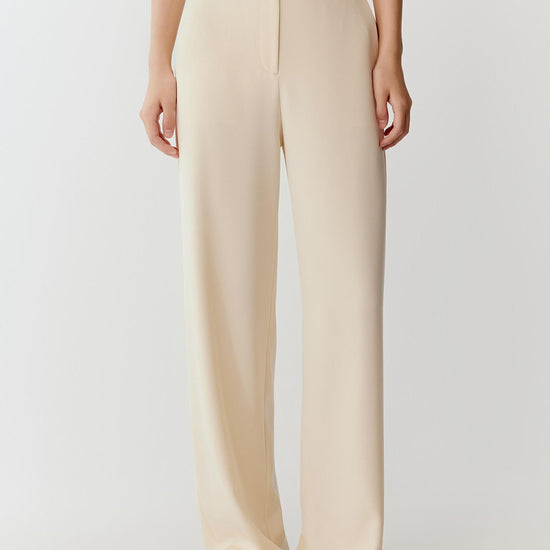 Woman wearing ivory silk stretch heavy crepe trousers with a fluid, wide-leg cut, styled with a sleeveless top. She moves through front and side views, showing the smooth fabric, soft structure and refined drape. The high waist and clean lines remain visible, while the trousers fall straight along the leg, emphasizing fluid movement and a relaxed silhouette.