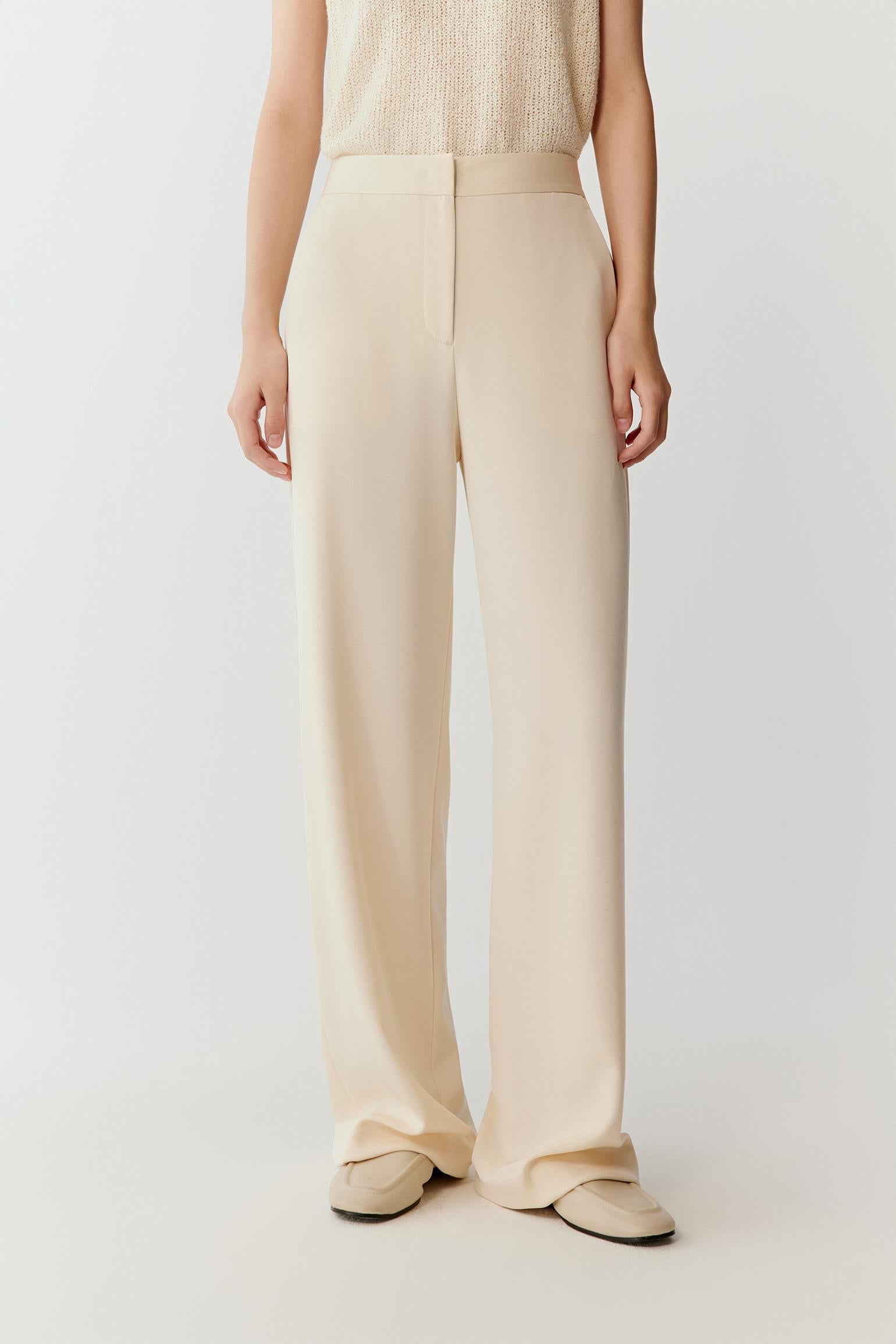 Woman wearing ivory silk stretch heavy crepe trousers with a fluid, wide-leg cut, styled with a sleeveless top. She moves through front and side views, showing the smooth fabric, soft structure and refined drape. The high waist and clean lines remain visible, while the trousers fall straight along the leg, emphasizing fluid movement and a relaxed silhouette.