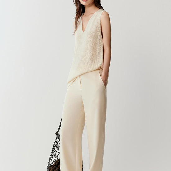 Woman wearing an ivory sleeveless silk knit top with a relaxed, fluid silhouette and V-neckline, styled with matching trousers. She moves through front, side and slight walking views, showing the lightweight drape, soft texture and natural movement of the knit. The fabric follows the body gently, highlighting the clean lines, open armholes and breathable construction, with subtle texture visible as the light shifts.