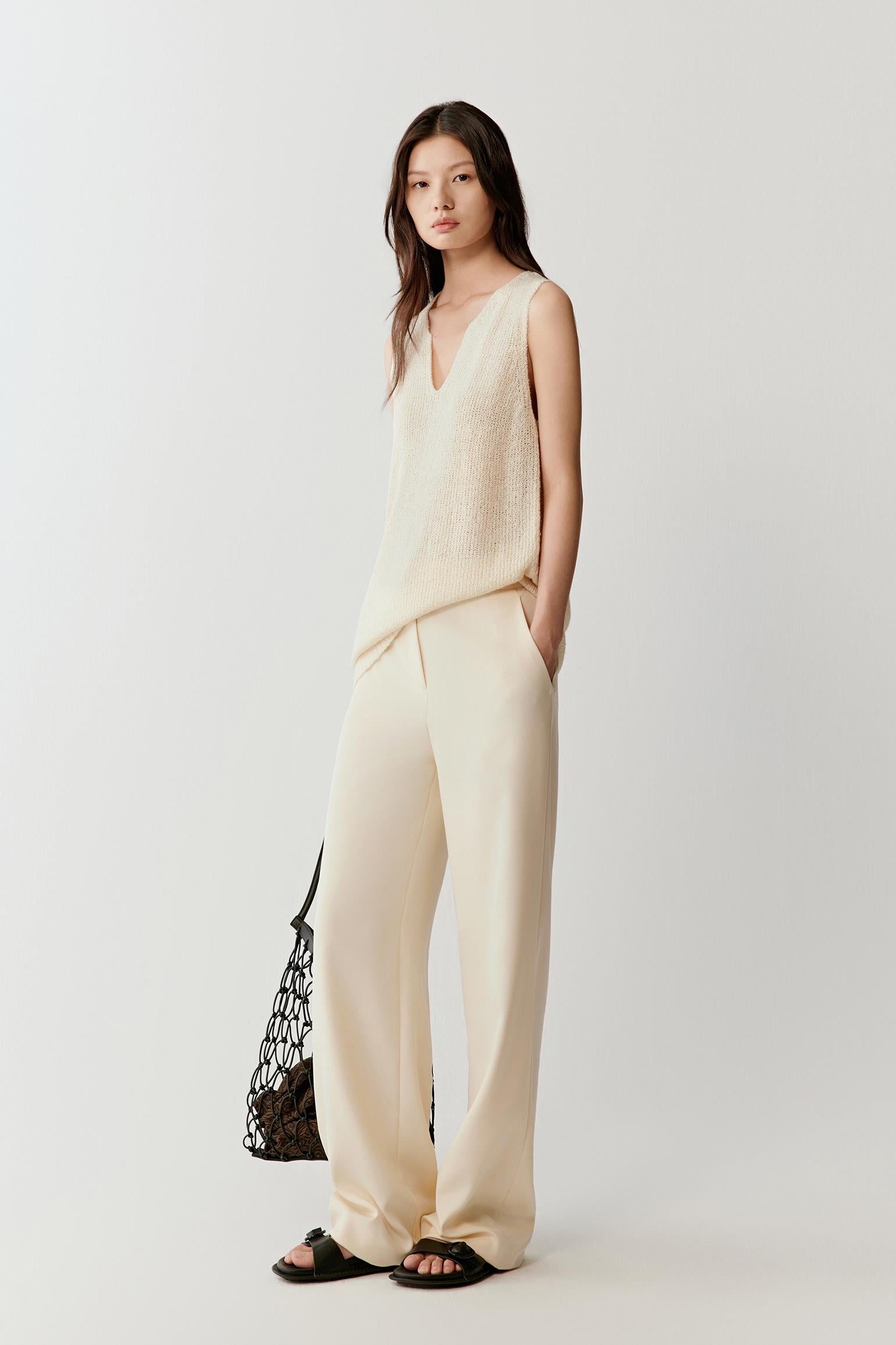Woman wearing an ivory sleeveless silk knit top with a relaxed, fluid silhouette and V-neckline, styled with matching trousers. She moves through front, side and slight walking views, showing the lightweight drape, soft texture and natural movement of the knit. The fabric follows the body gently, highlighting the clean lines, open armholes and breathable construction, with subtle texture visible as the light shifts.