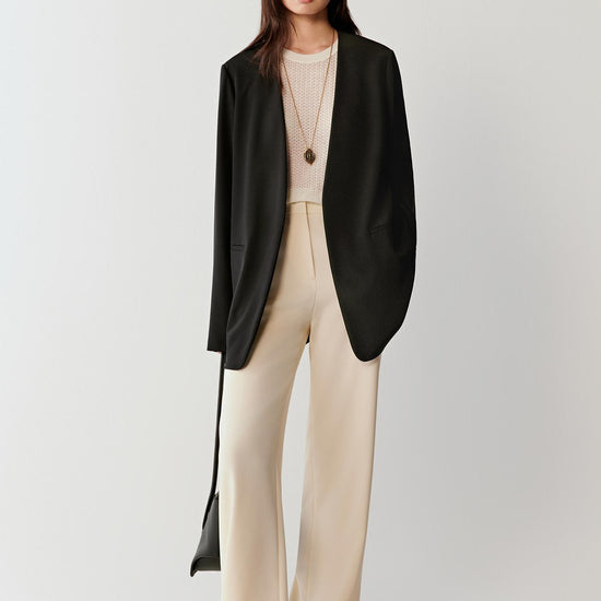 Woman wearing a black silk stretch crepe blazer with a straight, relaxed cut, styled with light beige trousers. She moves through front, three-quarter and side views, showing the fluid drape, soft shoulders and lightweight structure. The blazer falls cleanly along the body, highlighting its straight silhouette, refined lapel and smooth, slightly matte texture, with natural movement emphasizing comfort and ease.