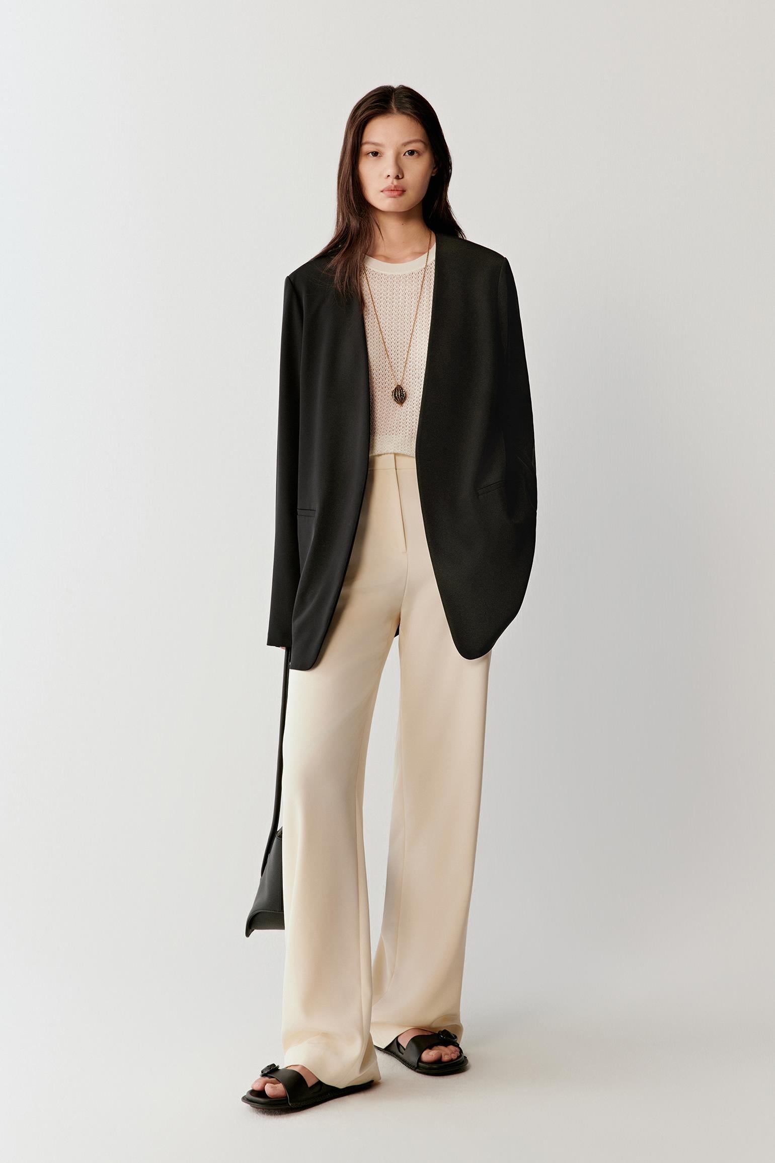 Woman wearing a black silk stretch crepe blazer with a straight, relaxed cut, styled with light beige trousers. She moves through front, three-quarter and side views, showing the fluid drape, soft shoulders and lightweight structure. The blazer falls cleanly along the body, highlighting its straight silhouette, refined lapel and smooth, slightly matte texture, with natural movement emphasizing comfort and ease.