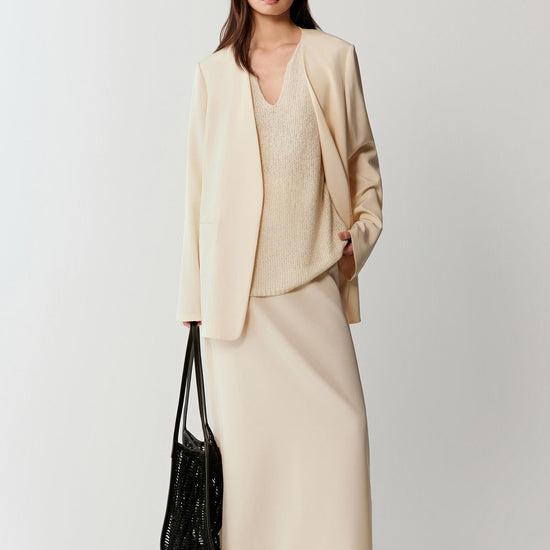 Woman wearing an ivory silk stretch crepe blazer with a straight, relaxed cut and fluid drape, styled with matching trousers. She moves from front to side and slight back angles, showing the clean lines, soft shoulders and lightweight structure. The fabric follows the body with a smooth, fluid movement, highlighting the length of the blazer, straight silhouette and refined lapel construction.