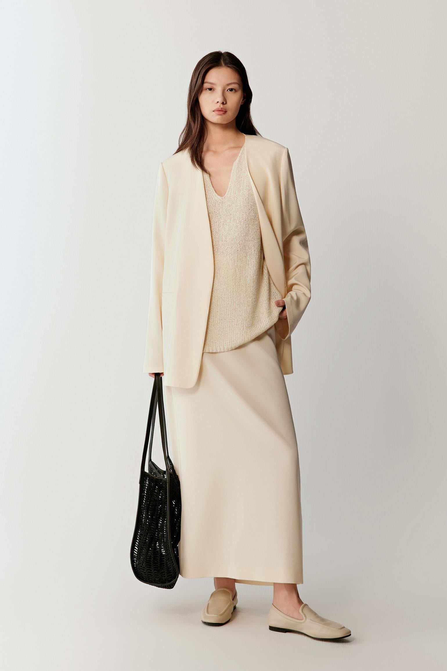 Woman wearing an ivory silk stretch crepe blazer with a straight, relaxed cut and fluid drape, styled with matching trousers. She moves from front to side and slight back angles, showing the clean lines, soft shoulders and lightweight structure. The fabric follows the body with a smooth, fluid movement, highlighting the length of the blazer, straight silhouette and refined lapel construction.