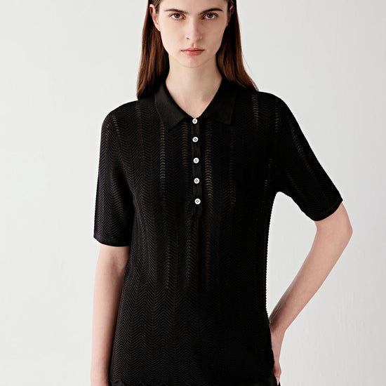 Video showing a woman wearing a deep black polo knit in silk and cotton, moving to reveal the fluid drape and openwork chevron knit structure. The lightweight knit develops subtle transparency and texture as the body moves, while the polo collar and front button placket remain structured and defined. The motion highlights the elongated silhouette, soft hand and breathable open knit construction, with the textured chevron surface catching light across the fabric.