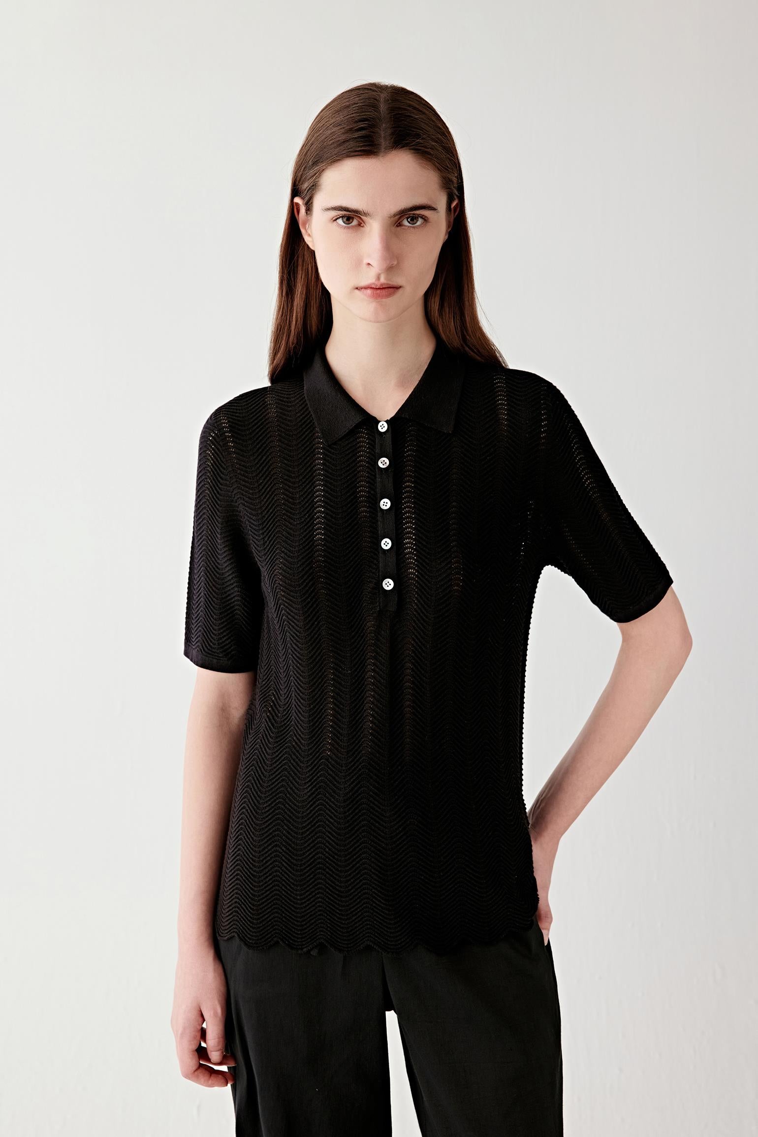 Video showing a woman wearing a deep black polo knit in silk and cotton, moving to reveal the fluid drape and openwork chevron knit structure. The lightweight knit develops subtle transparency and texture as the body moves, while the polo collar and front button placket remain structured and defined. The motion highlights the elongated silhouette, soft hand and breathable open knit construction, with the textured chevron surface catching light across the fabric.