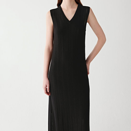 Video showing a woman wearing a deep black maxi dress in silk and cotton knit, moving to reveal the fluid drape and openwork chevron knit structure. The lightweight knit develops subtle transparency and soft movement through the length of the silhouette. The V-neckline and flared hem remain defined while the fabric flows naturally with motion, highlighting the airy construction and textured knit surface.