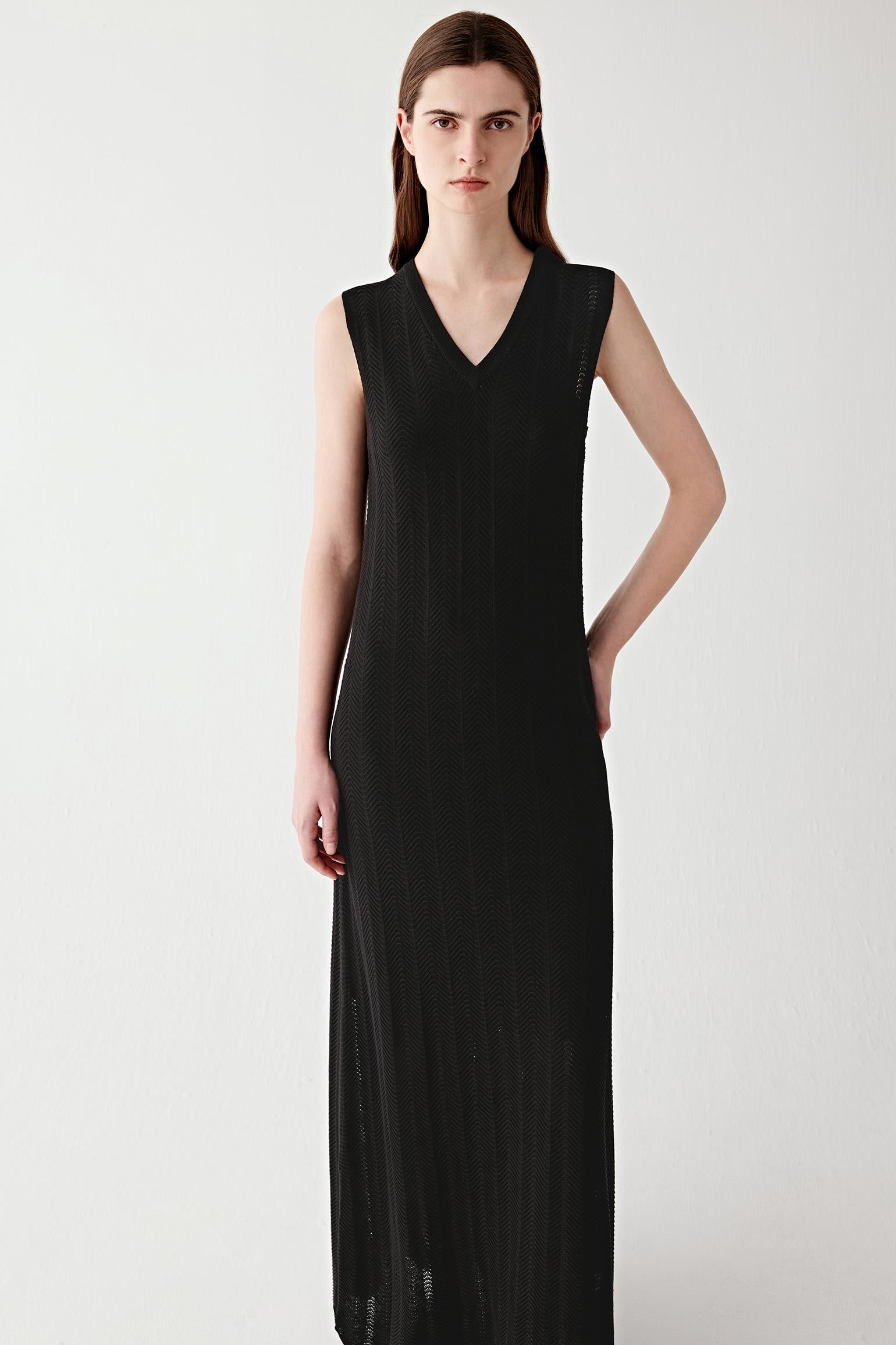 Video showing a woman wearing a deep black maxi dress in silk and cotton knit, moving to reveal the fluid drape and openwork chevron knit structure. The lightweight knit develops subtle transparency and soft movement through the length of the silhouette. The V-neckline and flared hem remain defined while the fabric flows naturally with motion, highlighting the airy construction and textured knit surface.