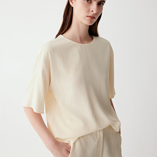 Video showing a woman wearing an off-white silk blouse with a softly crinkled surface and fluid drape, moving to reveal the lightweight texture and continuous silhouette. The fabric falls softly through the body and sleeves, while the side slit and back slit fastening open subtly with movement. The motion highlights the matte crinkled texture, relaxed structure and fluid construction, with the lightweight silk shifting naturally around the body.