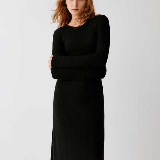 Woman wearing a long black fitted dress with long sleeves and black loafers.