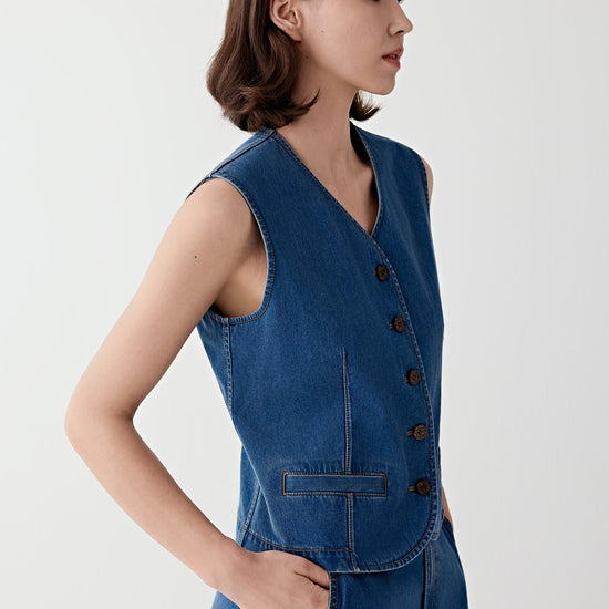 Woman wearing a blue cotton stretch denim vest with a fitted, sleeveless cut, styled with matching denim trousers. She moves through front and side views, showing the structured yet flexible fabric and clean, tailored lines. The V-neckline, button fastening and defined seams remain visible, while the vest follows the torso closely, emphasizing structure, ease of movement and a sharp silhouette.