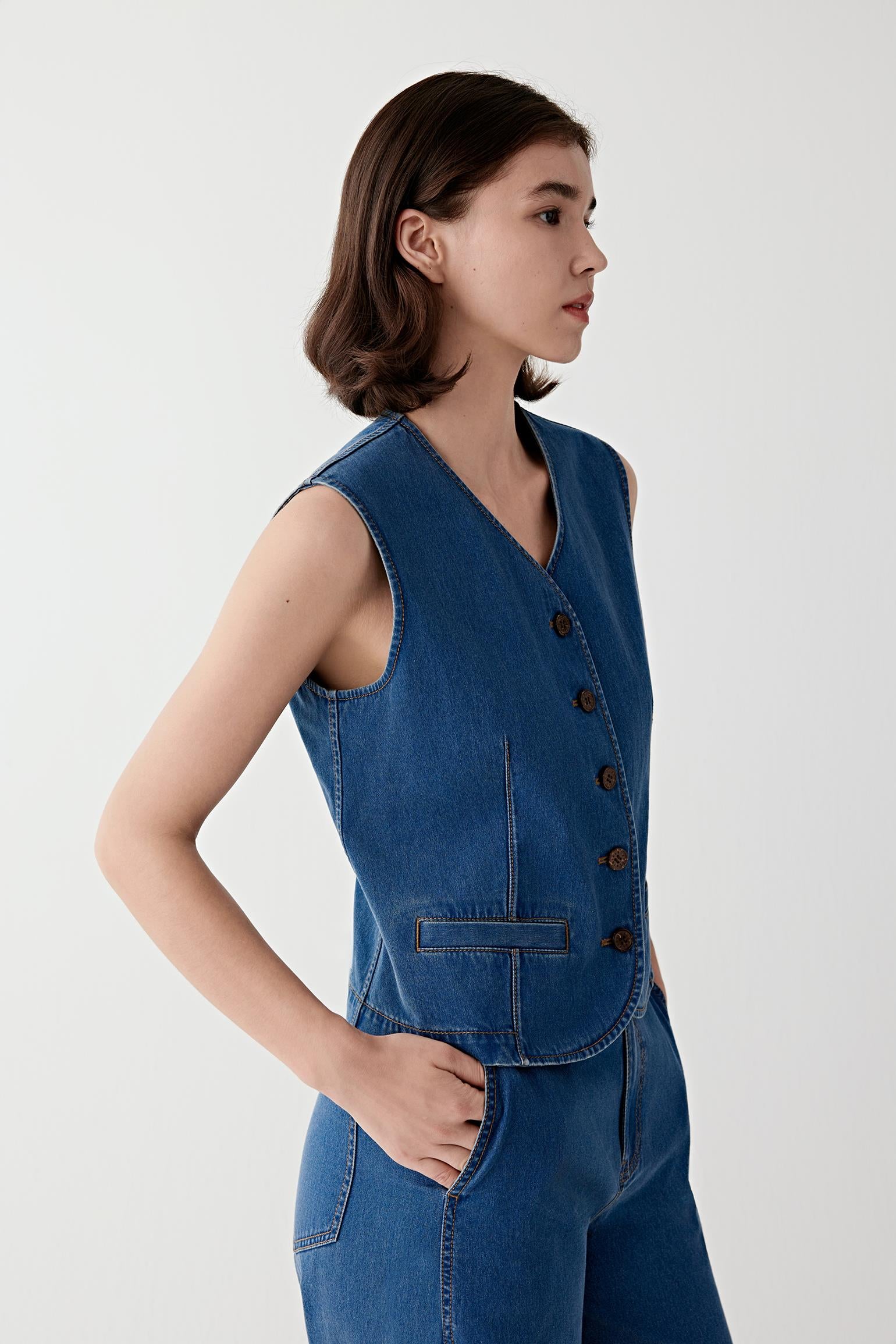 Woman wearing a blue cotton stretch denim vest with a fitted, sleeveless cut, styled with matching denim trousers. She moves through front and side views, showing the structured yet flexible fabric and clean, tailored lines. The V-neckline, button fastening and defined seams remain visible, while the vest follows the torso closely, emphasizing structure, ease of movement and a sharp silhouette.