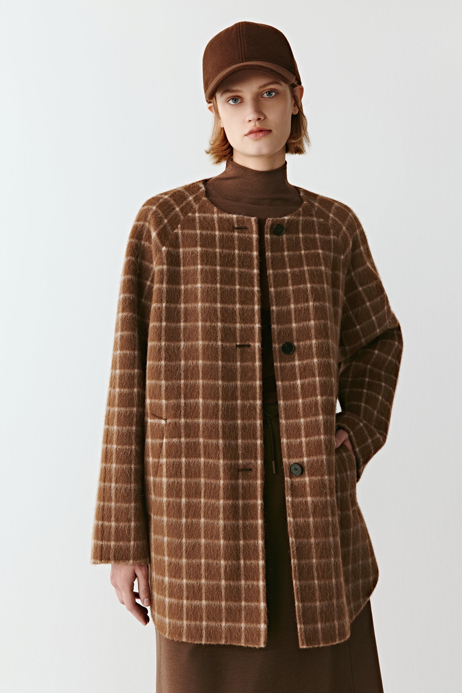 Product video showing a short double-faced checked wool coat in natural brown tones, rotating to display the check pattern, collarless round neckline, raglan sleeves, welt pockets, and overall silhouette from all angles.