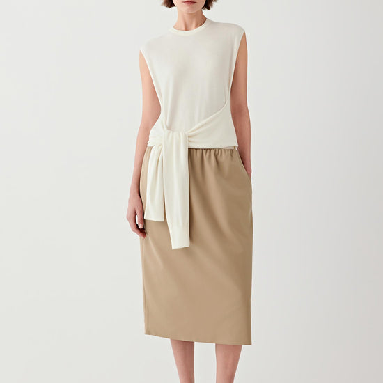 Video showing the model wearing the cream sleeveless pullover knitted from worsted wool while walking and turning. The sequence highlights the clean architectural silhouette of the garment, the sharply cut arm openings that frame the shoulders and the soft movement of the knit fabric around the waist. The front tie detail moves gently with the body, creating subtle volume and shaping.