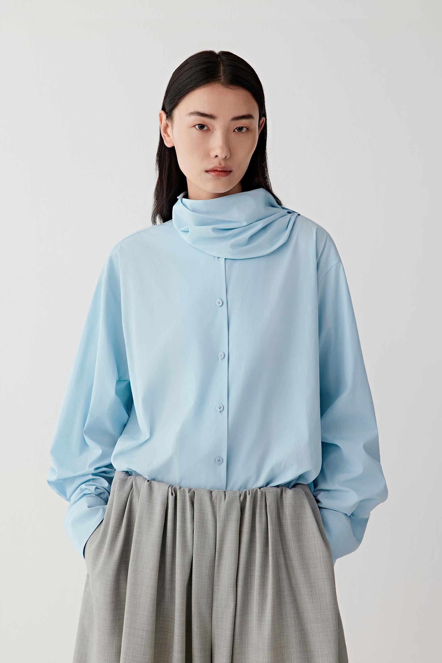 Video showing the model turning while wearing the light blue shirt, revealing the soft movement of the cotton poplin fabric and the sculptural fold of the collar.