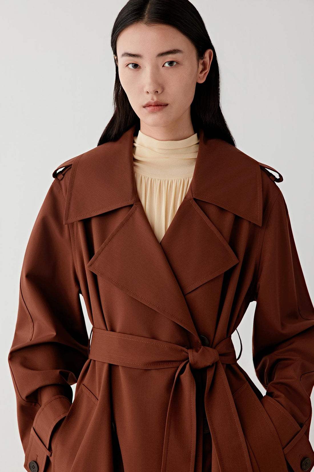 Video showing a woman wearing a long brown wool and silk trench coat with a wide collar and belt at the waist. The model moves slightly to show the drape and movement of the fabric in a studio setting.