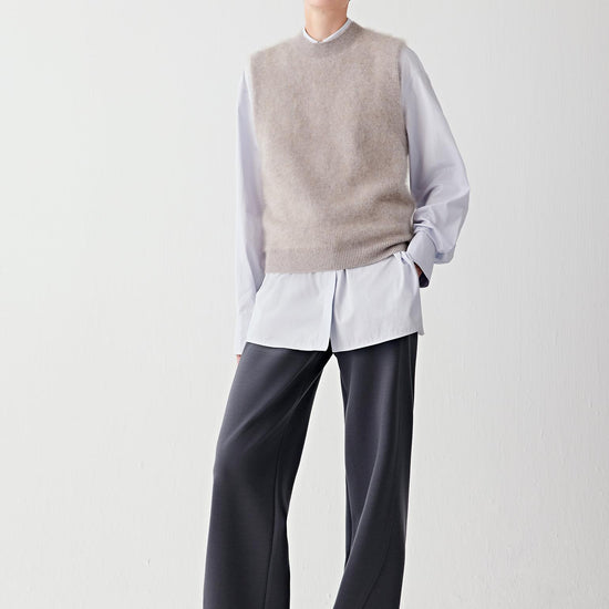 Video of a woman standing against a plain light background, wearing a light grey sleeveless knit over a white shirt, paired with dark straight-leg trousers and beige textured shoes.