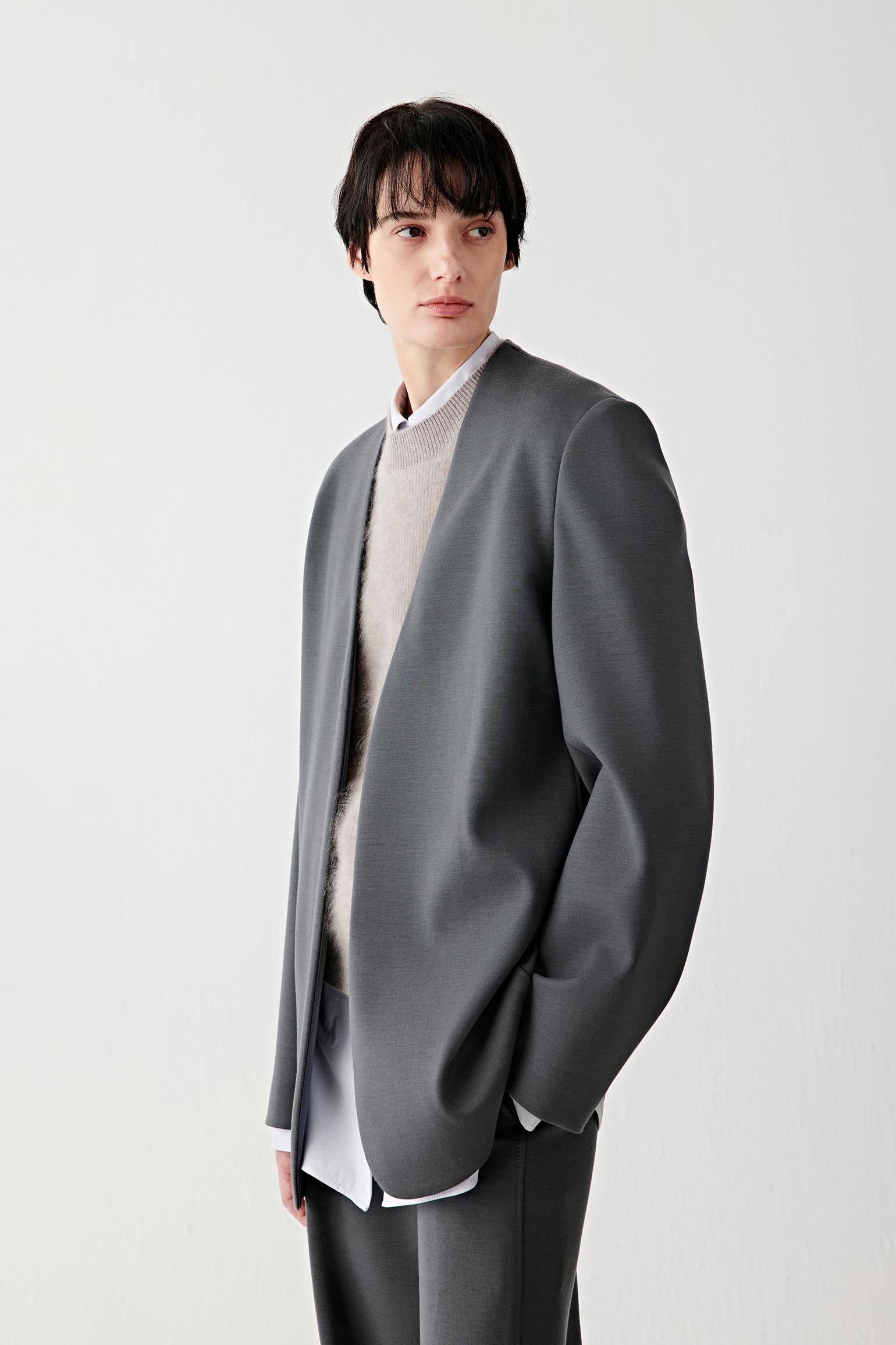 Video of a woman standing against a plain light background, wearing a dark grey collarless jacket over a light grey knit and white shirt, paired with matching straight-leg trousers and beige textured shoes.