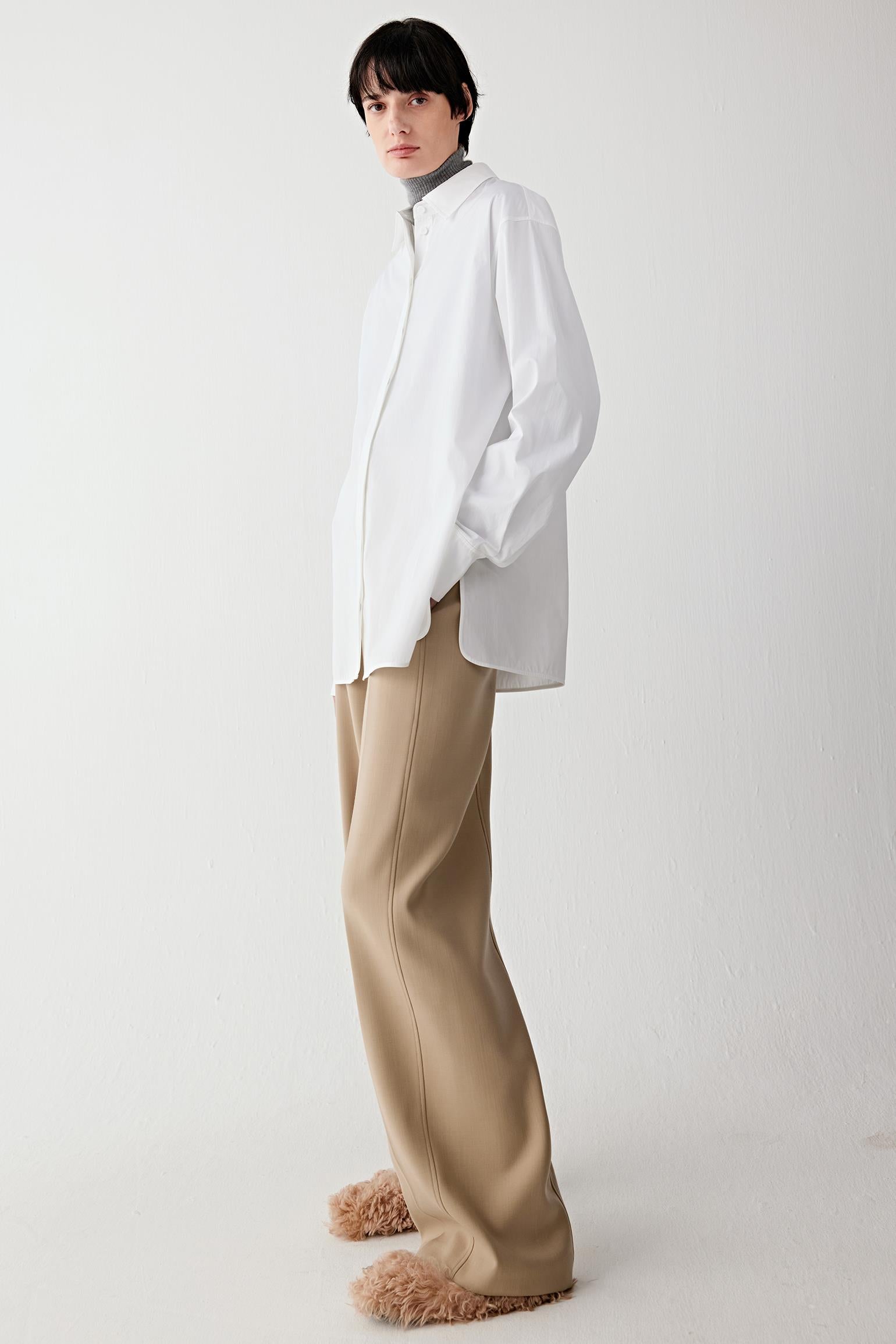 Video of a woman standing against a plain light background, wearing an oversized white shirt layered over a grey turtleneck, paired with camel straight-leg trousers and beige textured shoes.