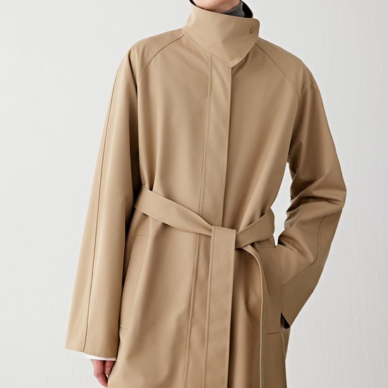 Video of a woman standing against a plain light background, wearing a long camel coat with snap buttons, layered over a grey high-neck top and white shirt, paired with matching wide-leg trousers and beige textured shoes.