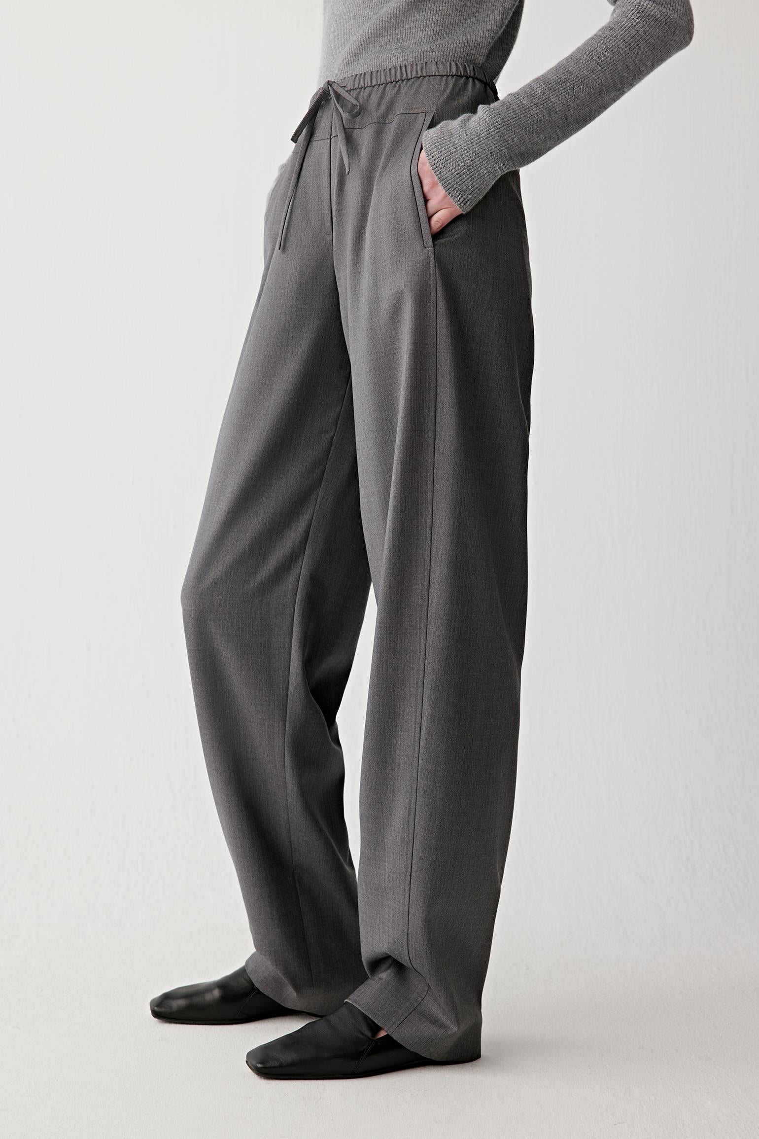 Video of a woman standing against a plain light background, wearing a fitted grey turtleneck sweater paired with dark grey drawstring trousers and black shoes.