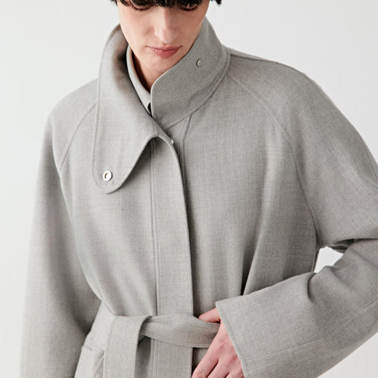 Woman wearing a light grey double-faced wool jacket with a relaxed, straight cut and self-tie belt, styled with matching trousers. She moves through front, side and back views, showing the soft, lightly structured fabric and fluid drape. The clean lines, wrap-style closure and belt detail remain visible, while the jacket falls naturally around the body, emphasizing comfort, ease of movement and a fluid silhouette.