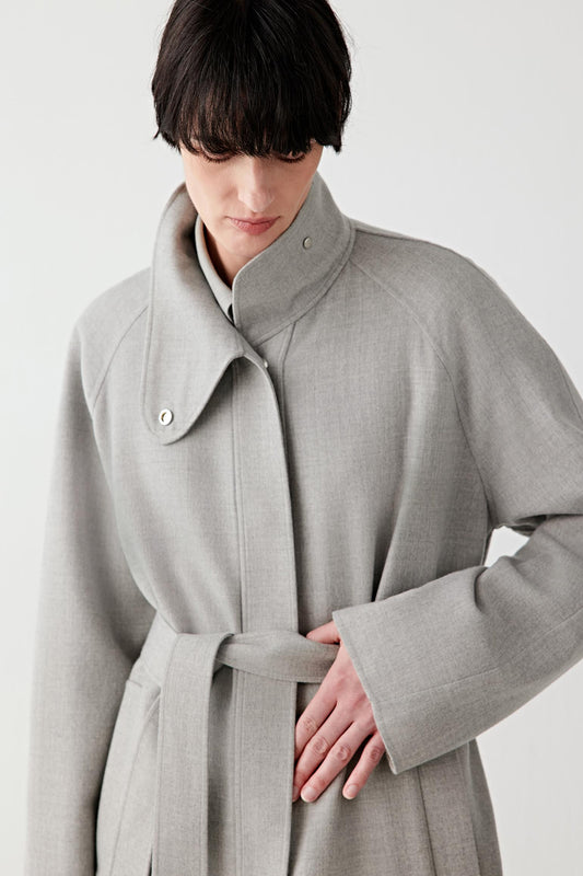 Woman wearing a light grey double-faced wool jacket with a relaxed, straight cut and self-tie belt, styled with matching trousers. She moves through front, side and back views, showing the soft, lightly structured fabric and fluid drape. The clean lines, wrap-style closure and belt detail remain visible, while the jacket falls naturally around the body, emphasizing comfort, ease of movement and a fluid silhouette.