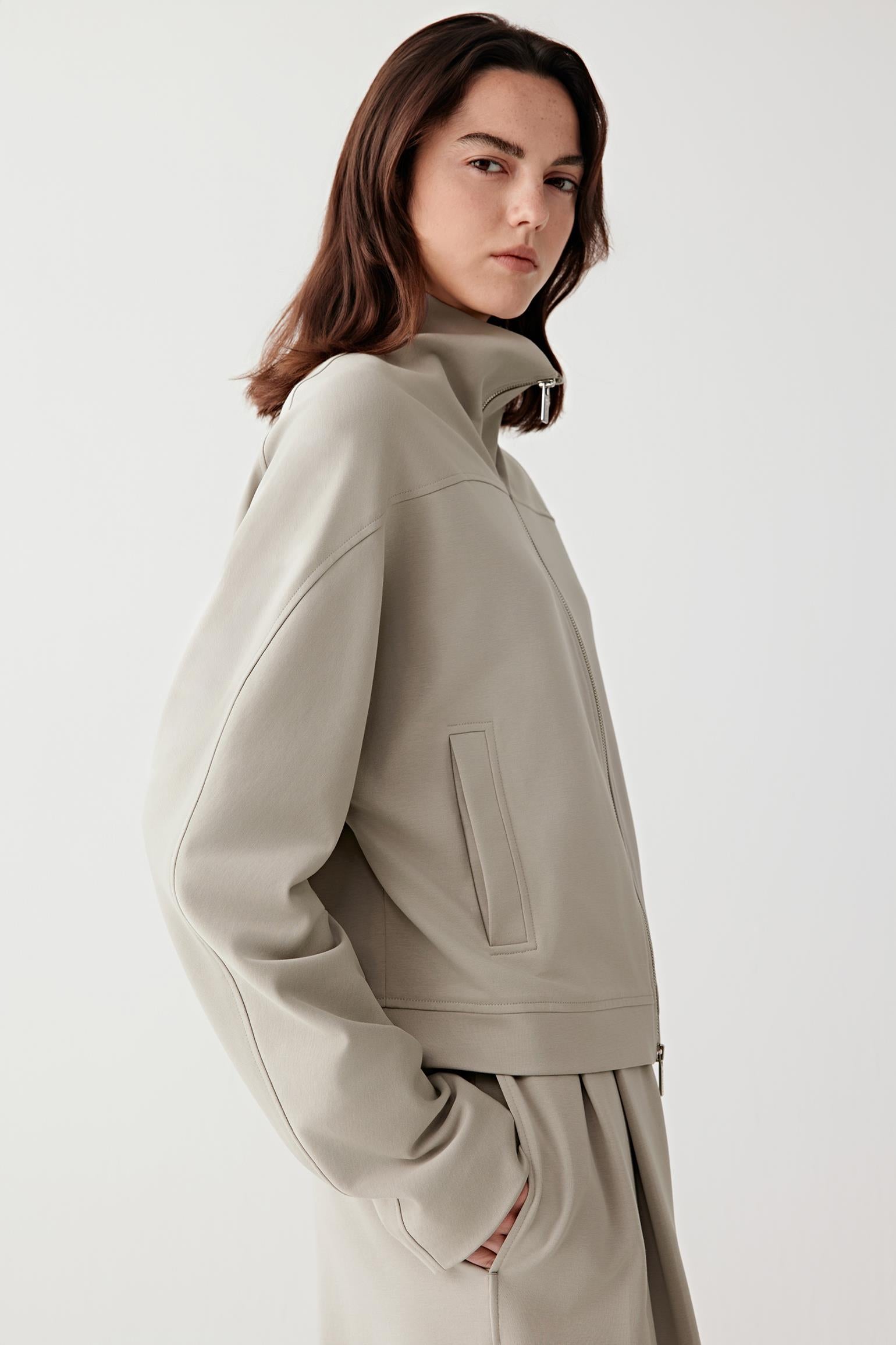 Video of a woman standing against a plain light background, wearing a light grey zip-up jacket with a high collar and matching wide-leg trousers, paired with beige textured shoes.