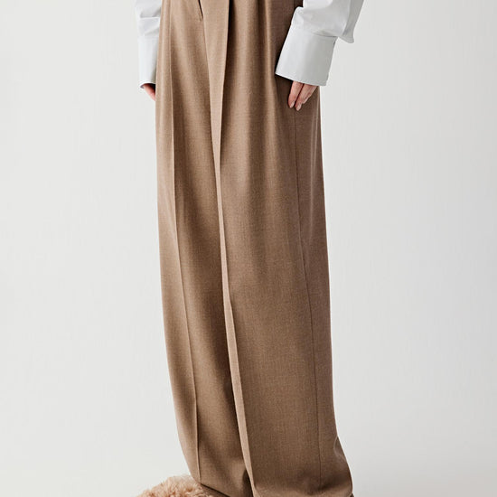 Video of a woman standing against a plain light background, wearing a light blue oversized shirt tucked into brown pleated wide-leg trousers, paired with beige textured shoes.