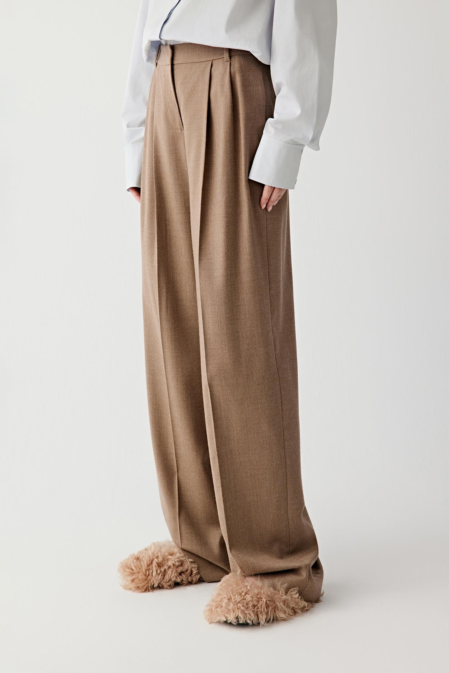 Video of a woman standing against a plain light background, wearing a light blue oversized shirt tucked into brown pleated wide-leg trousers, paired with beige textured shoes.