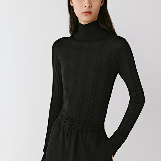 A woman wears a black ribbed pullover in wool and silk, styled with wide black trousers. She turns gracefully, allowing the camera to capture the pullover’s fluid drape and fitted shape. The soft texture of the fabric and the purity of the lines are accentuated through gentle movement. The sequence moves between full-length and close-up views, revealing the elegance and timelessness of the piece.