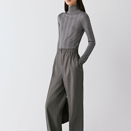 A woman wears grey melange straight-leg trousers in worsted wool, styled with a matching ribbed pullover. The video follows her as she walks slowly, showing the trousers from different angles. The light movement reveals the soft drape of the wool and the sharp pressed crease that defines the silhouette. The camera alternates between wider shots that capture the full look and closer frames focusing on the waistband and texture of the fabric, highlighting the comfort and refinement of the design.