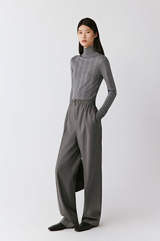 A woman wears grey melange straight-leg trousers in worsted wool, styled with a matching ribbed pullover. The video follows her as she walks slowly, showing the trousers from different angles. The light movement reveals the soft drape of the wool and the sharp pressed crease that defines the silhouette. The camera alternates between wider shots that capture the full look and closer frames focusing on the waistband and texture of the fabric, highlighting the comfort and refinement of the design.