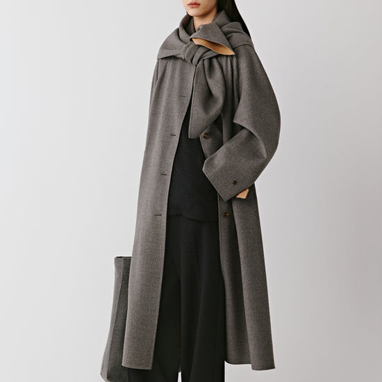 A woman wears a long grey double-faced wool coat styled with black trousers and boots. The video follows her as she moves calmly, revealing the coat from different angles. The camera alternates between wider shots that capture the full silhouette and closer views that linger on the collar, buttons and fabric texture. The natural drape of the wool and the coat’s balanced proportions are highlighted through gentle, continuous motion.