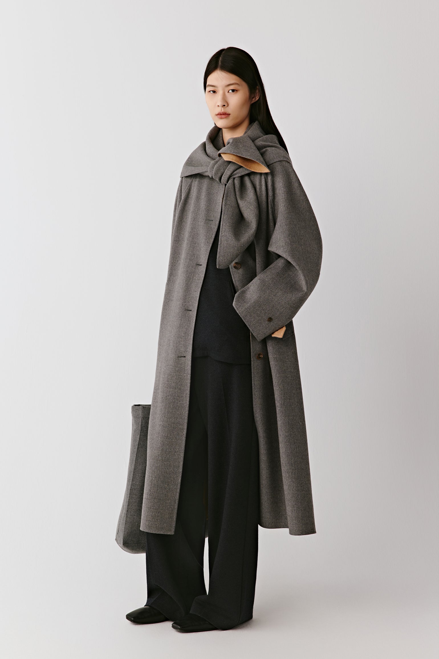 A woman wears a long grey double-faced wool coat styled with black trousers and boots. The video follows her as she moves calmly, revealing the coat from different angles. The camera alternates between wider shots that capture the full silhouette and closer views that linger on the collar, buttons and fabric texture. The natural drape of the wool and the coat’s balanced proportions are highlighted through gentle, continuous motion.