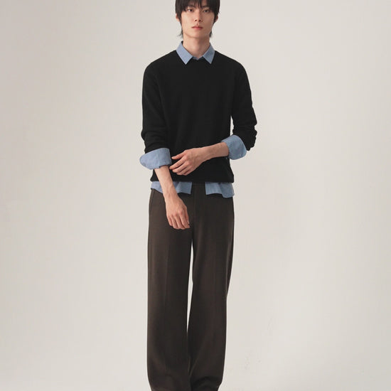 A man modeling a black long-sleeve knit sweater layered over a light blue collared shirt, paired with brown trousers and black shoes, against a plain light gray studio background.