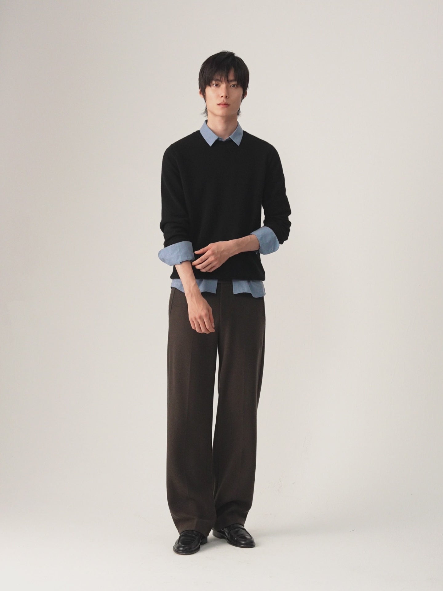A man modeling a black long-sleeve knit sweater layered over a light blue collared shirt, paired with brown trousers and black shoes, against a plain light gray studio background.